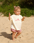 Child in a white shirt with text and pink shorts standing on sandy ground with greenery in the background