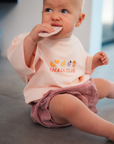 Baby wearing a pink 'Snack Club' shirt and plaid shorts, sitting on a floor.