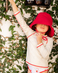 Child wearing a white outfit with red trim and a red hat against a leafy background