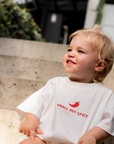 Child wearing a white t-shirt with 'Small But Spicy' text and red chili pepper graphic, sitting outdoors.