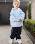 Child wearing a light blue hoodie and dark pants standing on a sidewalk.