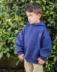 Child wearing a blue hoodie and olive pants standing in front of green foliage.