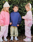 Three children in matching pink and blue outfits standing in front of a green bush.