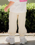Person wearing a white shirt with pink text, beige pants, and white shoes standing on a sidewalk with green bushes in the background.