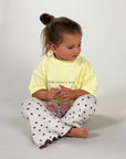 A toddler girl sitting cross-legged on a light gray studio floor, wearing a pale yellow graphic T-shirt with the text 'RUN NEEDS A MARGI' and white pants with a black star pattern.