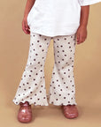 A child wearing cream flared pants with a dark brown star pattern, a white t-shirt, and pink shoes, standing against a solid beige background.