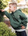 Child wearing a green hoodie with a star design, standing outdoors near greenery.