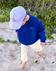 A toddler walking barefoot on sand near green bushes, wearing a royal blue sweatshirt, cream pants, and a light lavender baseball cap.