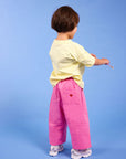 A toddler standing with their back to the camera, wearing a pale yellow t-shirt, bright pink wide-leg pants with a small red heart on the pocket, and white sneakers, set against a solid blue studio background.