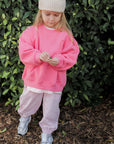 Child wearing a pink sweatshirt and light-colored pants standing in front of green foliage.