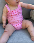 Baby wearing a pink playsuit with red strawberries sitting on a grey couch.