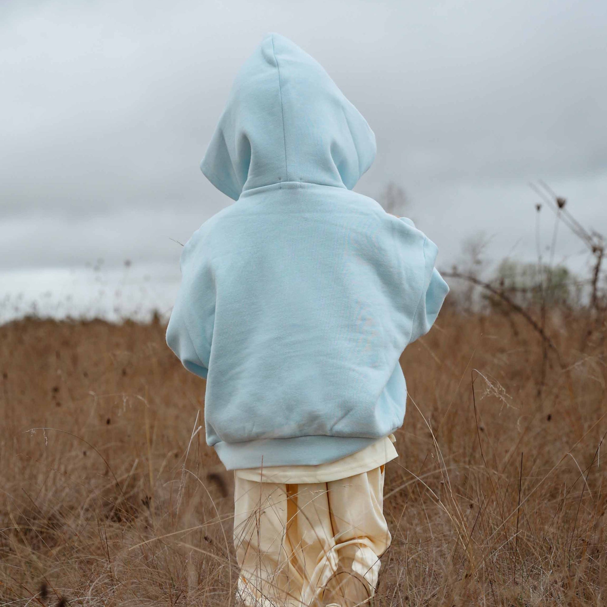 Person wearing a light blue hoodie and beige pants standing in a field with a cloudy sky.