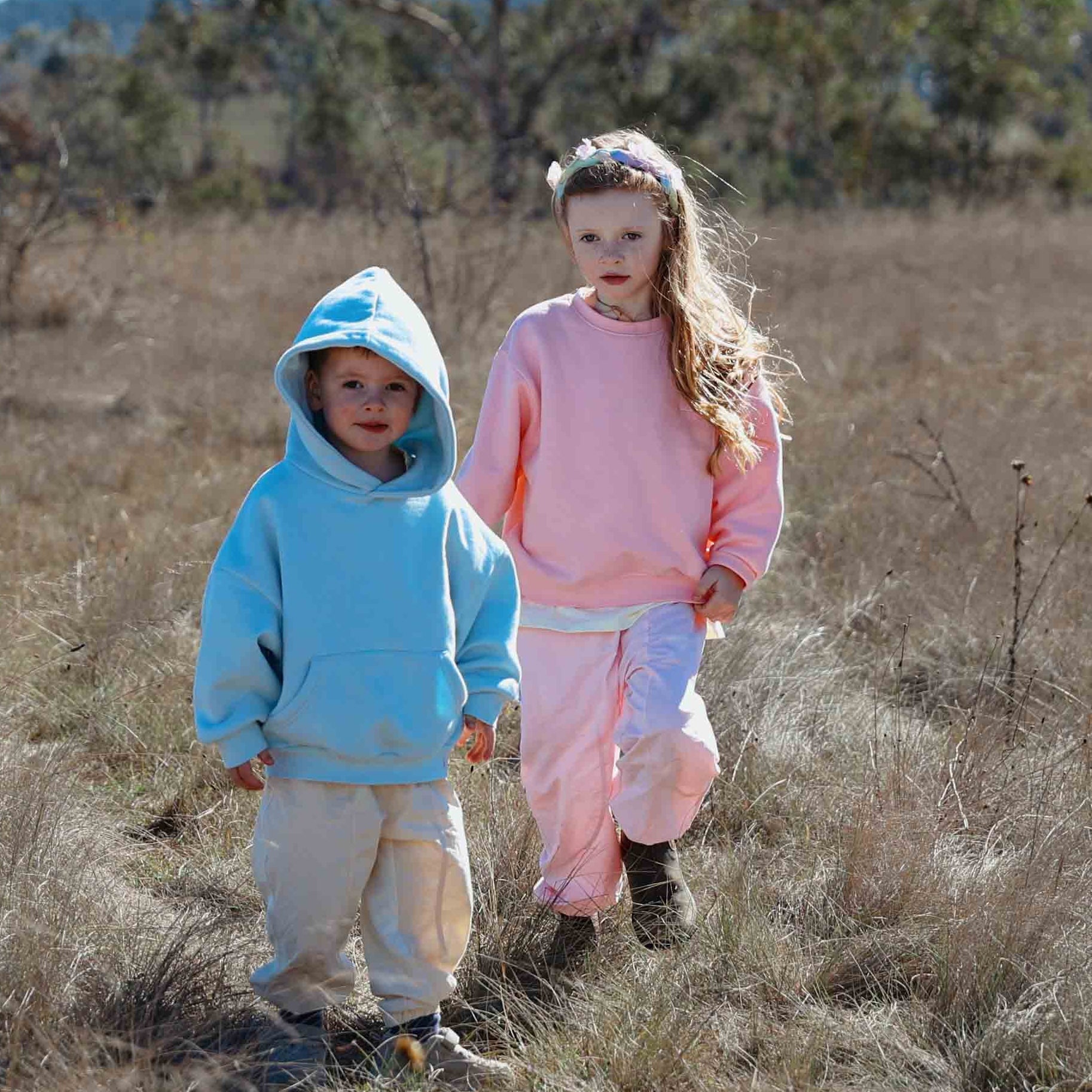 Two children in colorful outfits walking through a dry, open field with trees in the background.