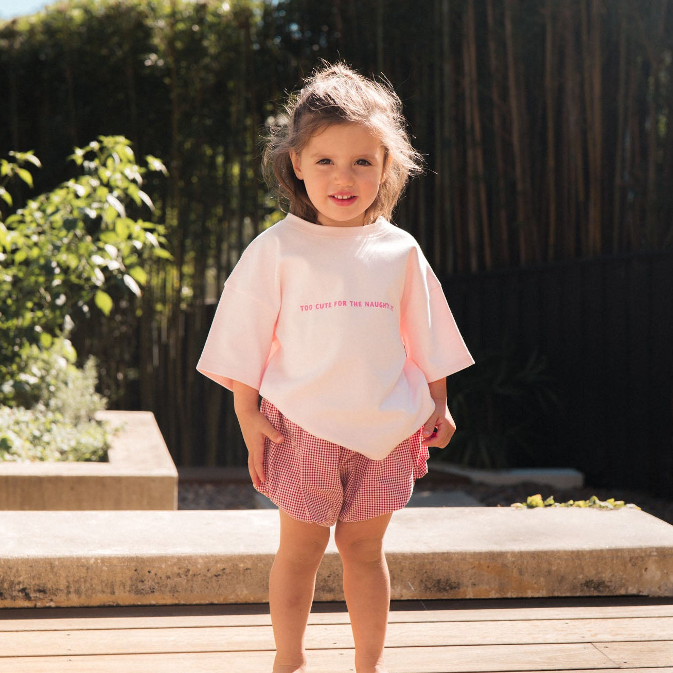Young girl in a pink outfit standing on a wooden deck with greenery in the background