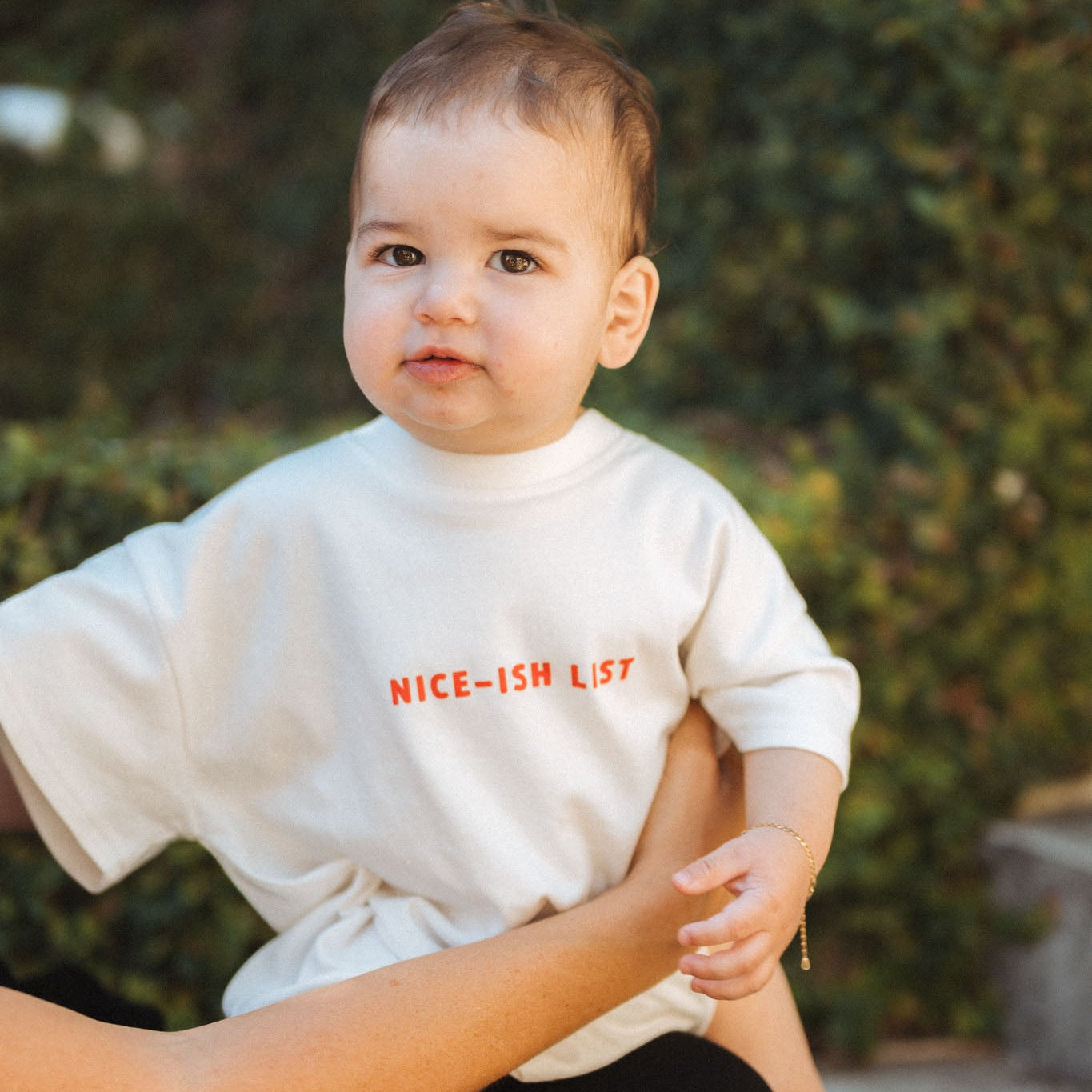 Baby wearing a white shirt with text, held by an adult outdoors.