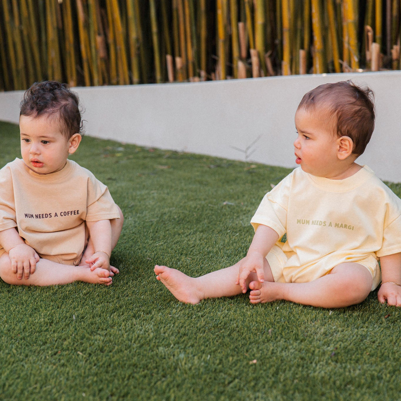 Two babies sitting on grass wearing matching outfits with text.