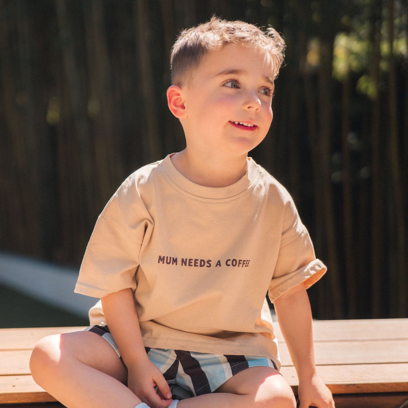 Child sitting on rocks wearing a beige t-shirt with text and checkered shorts.