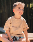 Child sitting on rocks wearing a beige t-shirt with text and checkered shorts.