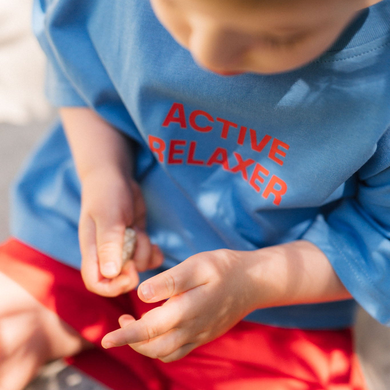 Child wearing a blue shirt with &#39;ACTIVE RELAXER&#39; text, holding small stones.