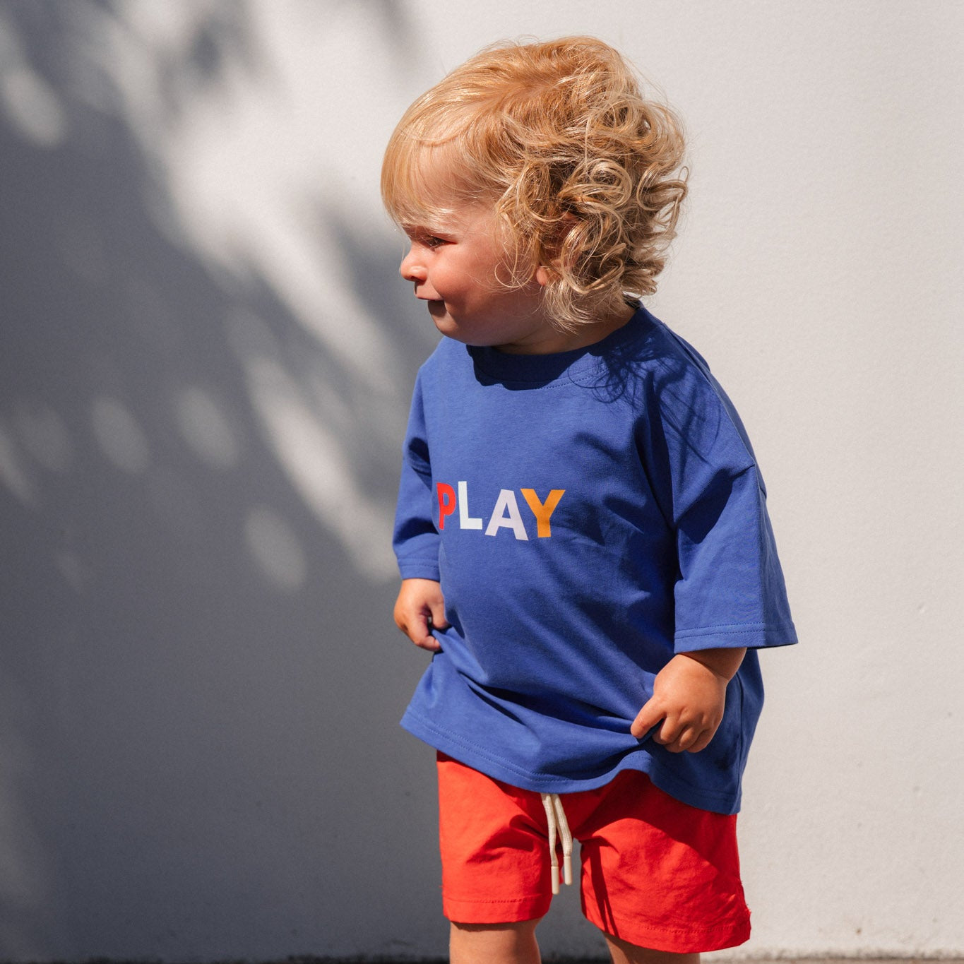 Child wearing a blue shirt with colorful letters and red shorts standing on a tiled floor.