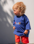 Child wearing a blue shirt with colorful letters and red shorts standing on a tiled floor.