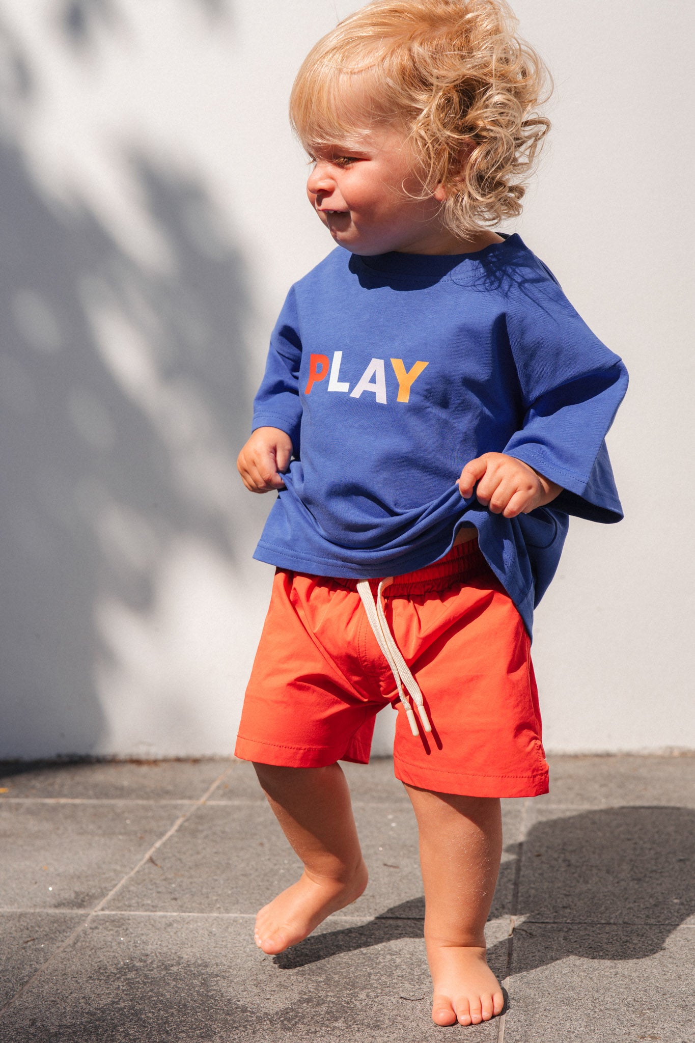 Child wearing a blue sweatshirt with 'PLAY' and red shorts standing on a gray surface.