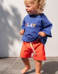 Child wearing a blue sweatshirt with 'PLAY' and red shorts standing on a gray surface.