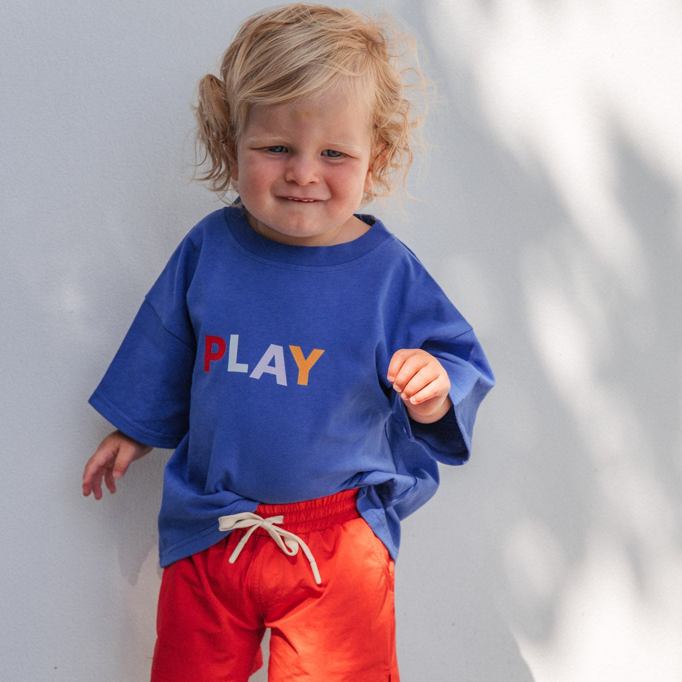 Child wearing a blue shirt with 'PLAY' and red shorts standing against a white wall.