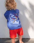 Child wearing a blue sleeveless shirt with a graphic design and red shorts against a white wall.