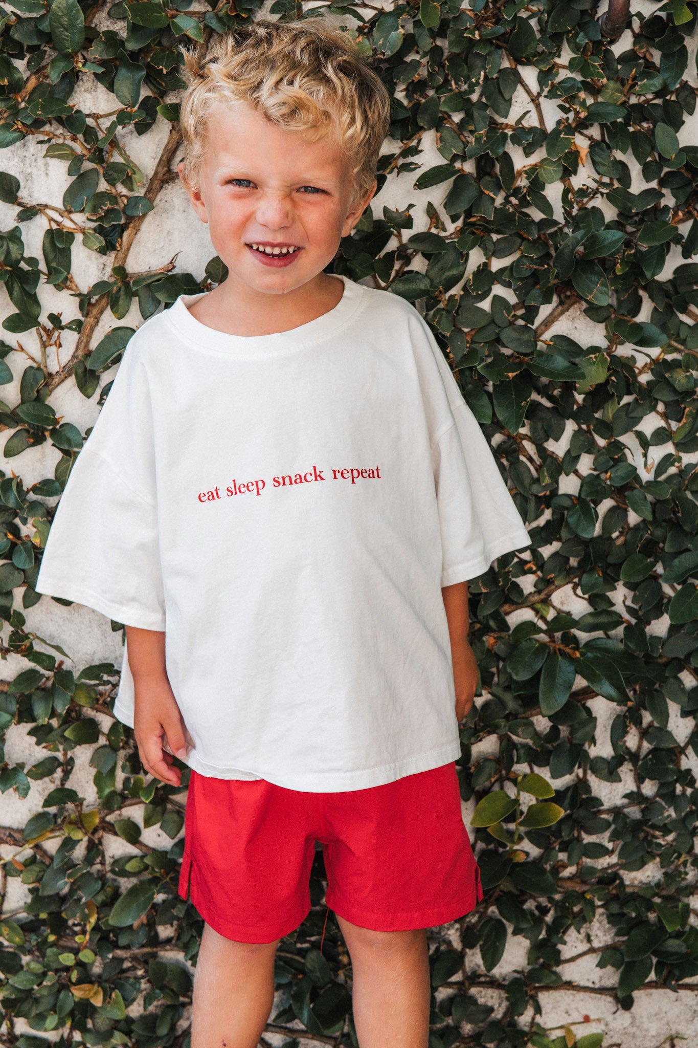 Child wearing a white t-shirt with eat sleep snack repeat and red shorts standing against a leafy background