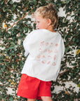 Child wearing a eat sleep snack repeat tee with favourite food patterns and red shorts standing against a leafy wall.