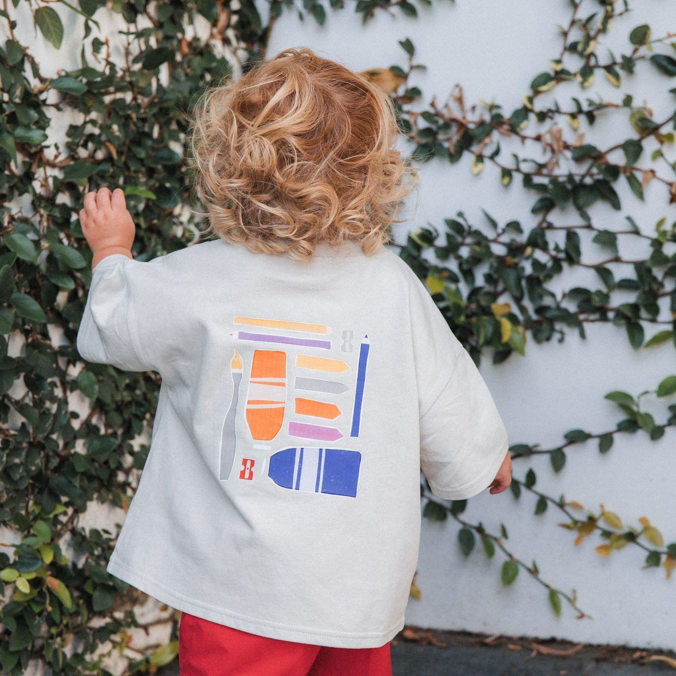 Child wearing a make more art tee with colorful graphics and red shorts, standing against a wall with greenery.