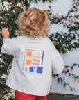 Child wearing a make more art tee with colorful graphics and red shorts, standing against a wall with greenery.
