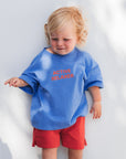 Child wearing a blue tee with active relaxer ext and red shorts standing against a white wall.