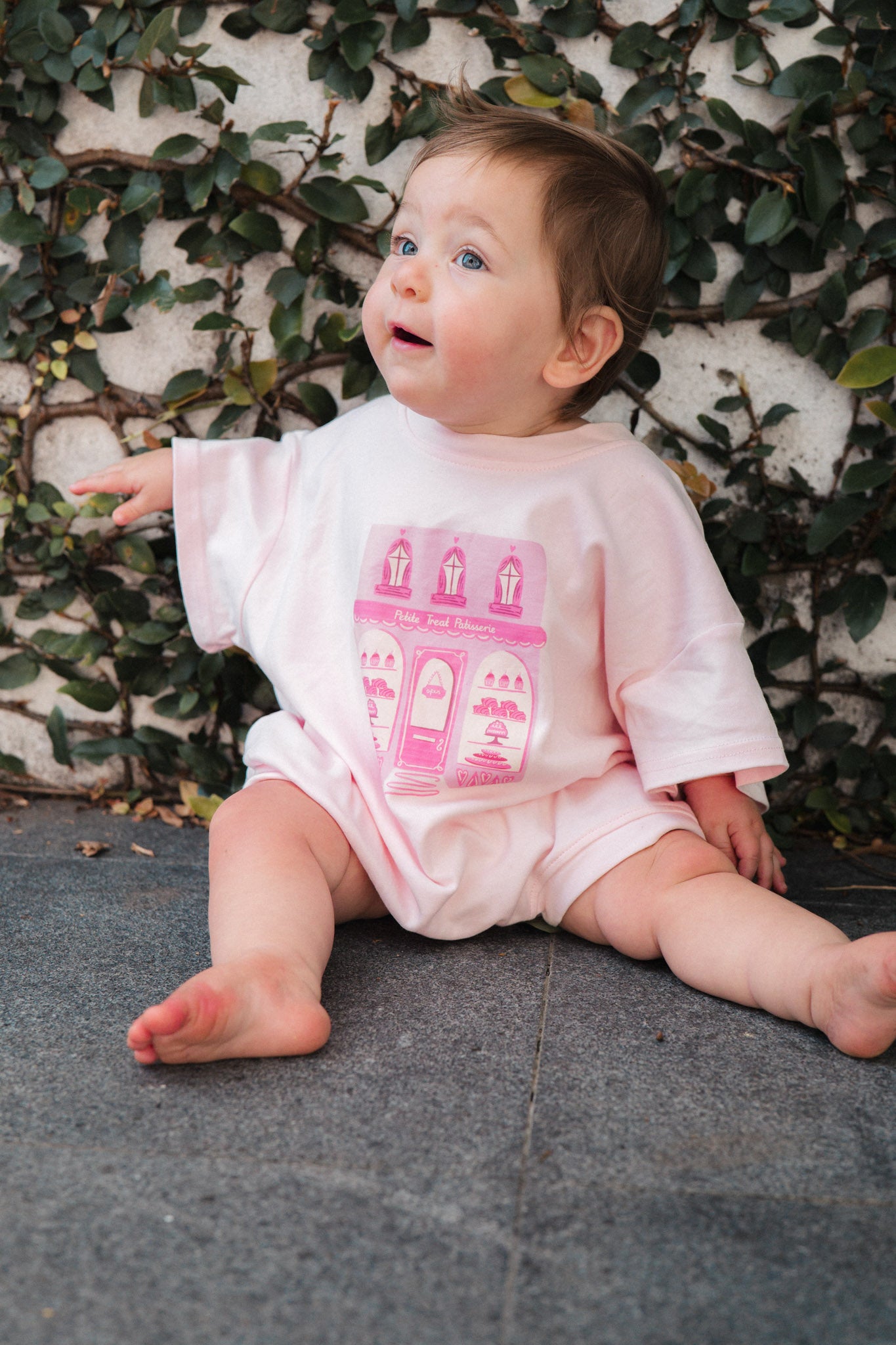 Baby wearing a pink patisserie bakery design, sitting on a stone surface with a leafy background.