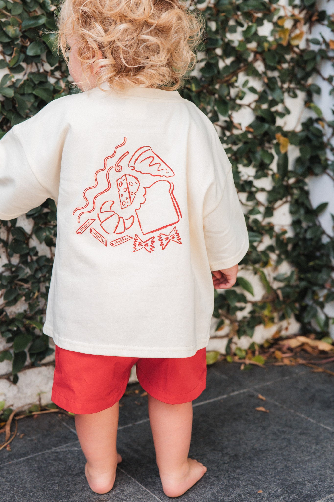 Child wearing a cream shirt with red graphics and red shorts standing in front of a green leafy background.