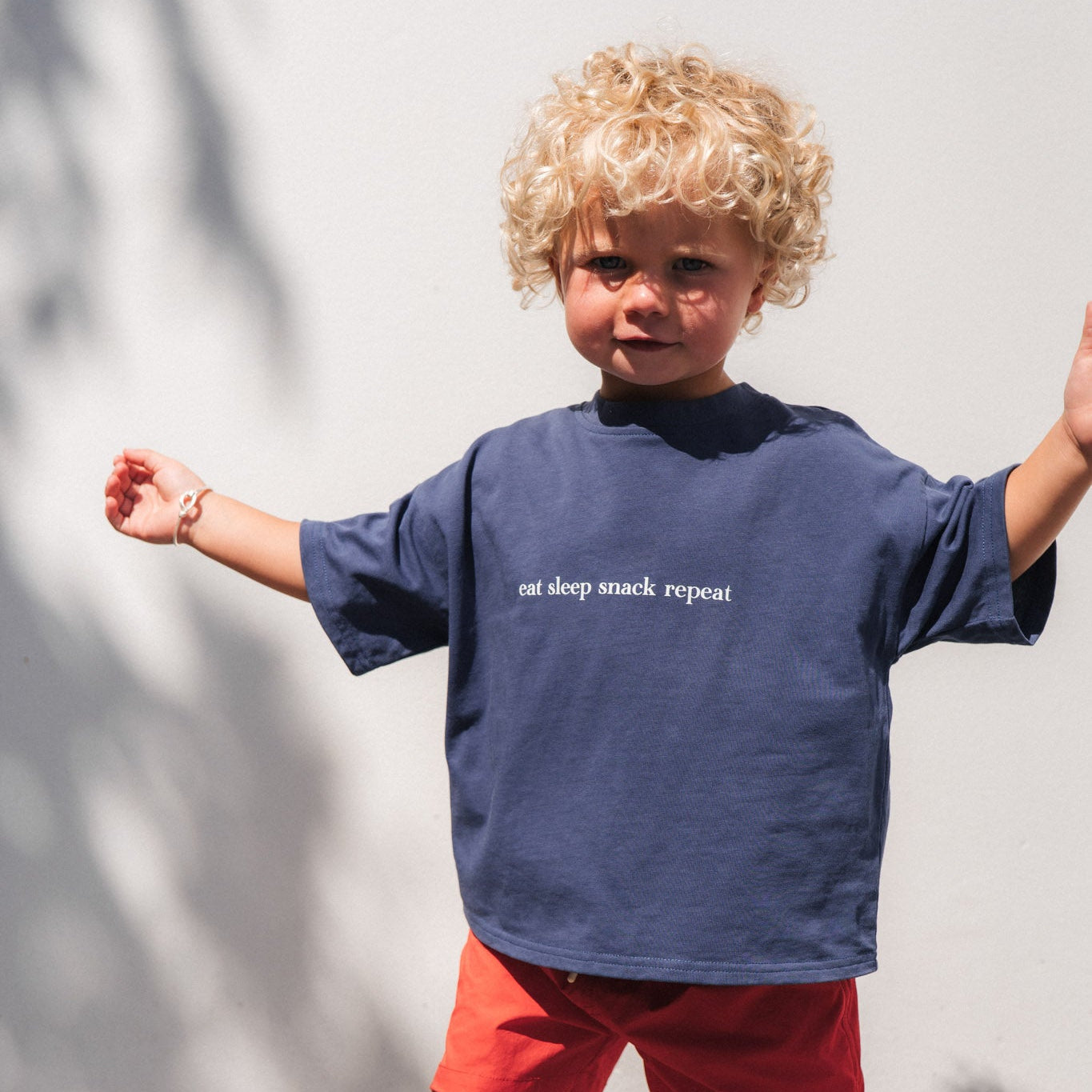 Child wearing a navy eat sleep snack repeat t-shirt with text and red shorts standing on a tiled floor.