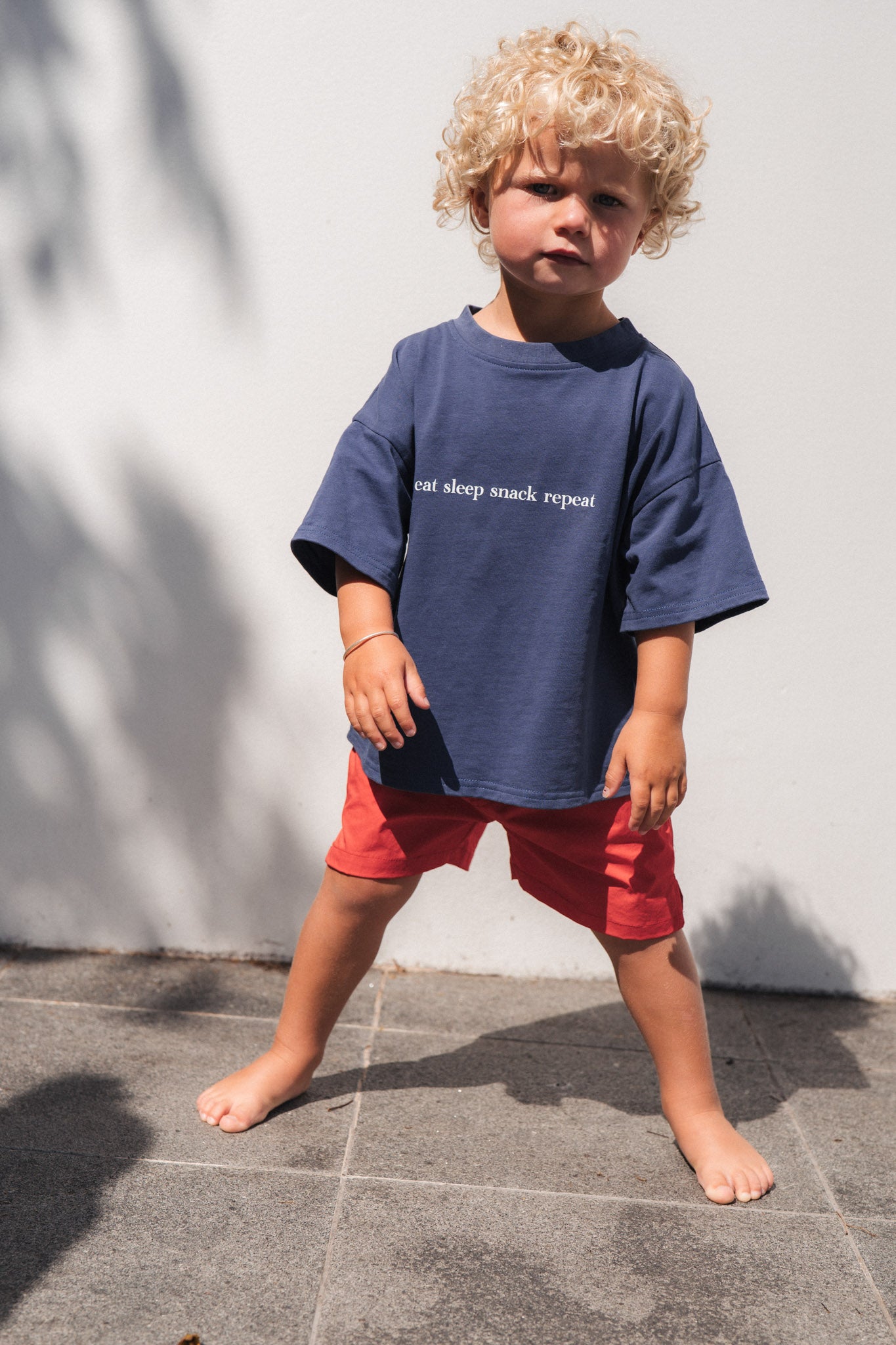 Child wearing eat sleep snack repeat shirt and red shorts standing against a white wall.