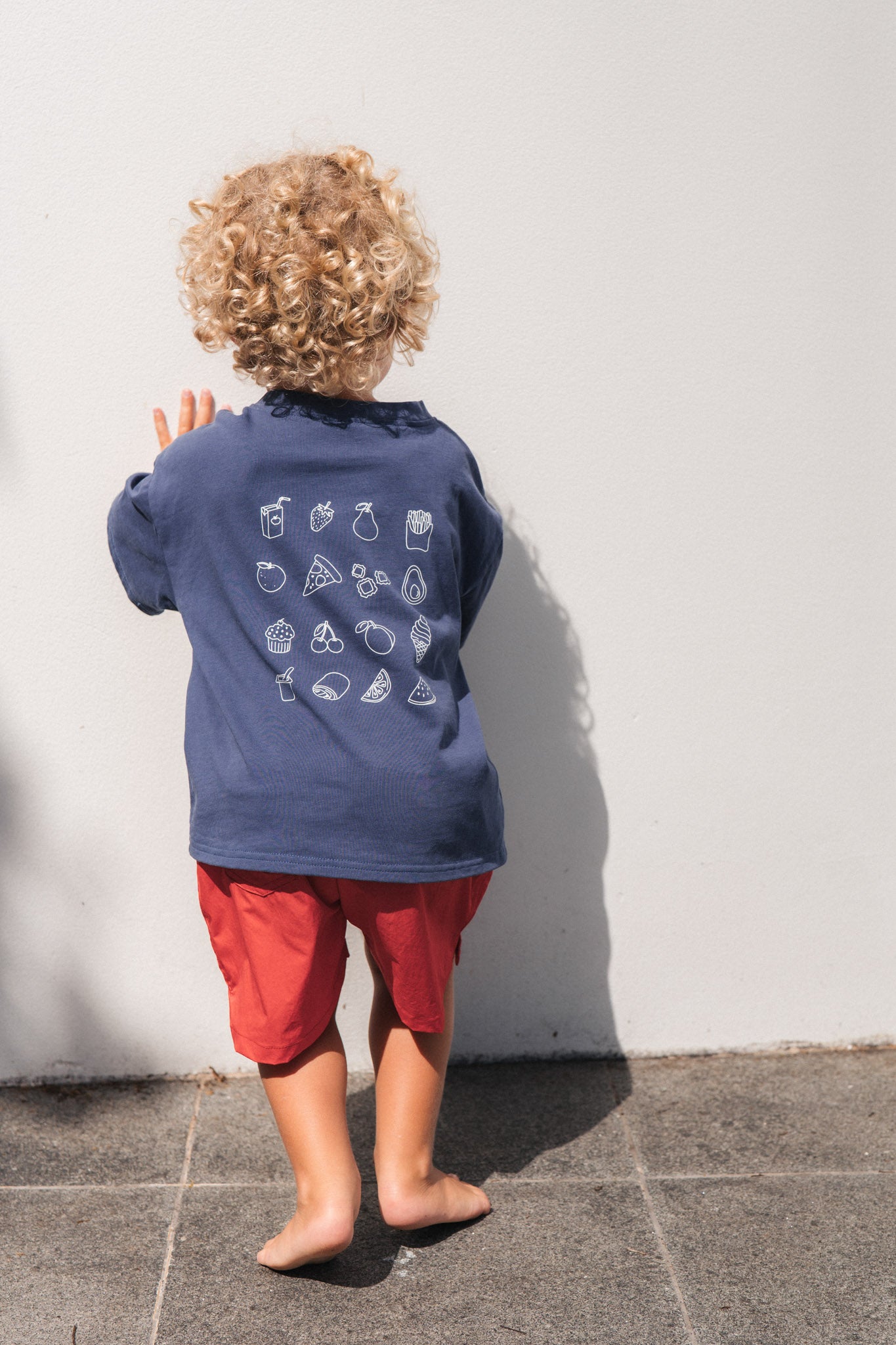 Child wearing eat sleep snack repeat tee with white graphics and red shorts standing against a white wall.