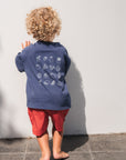 Child wearing eat sleep snack repeat tee with white graphics and red shorts standing against a white wall.