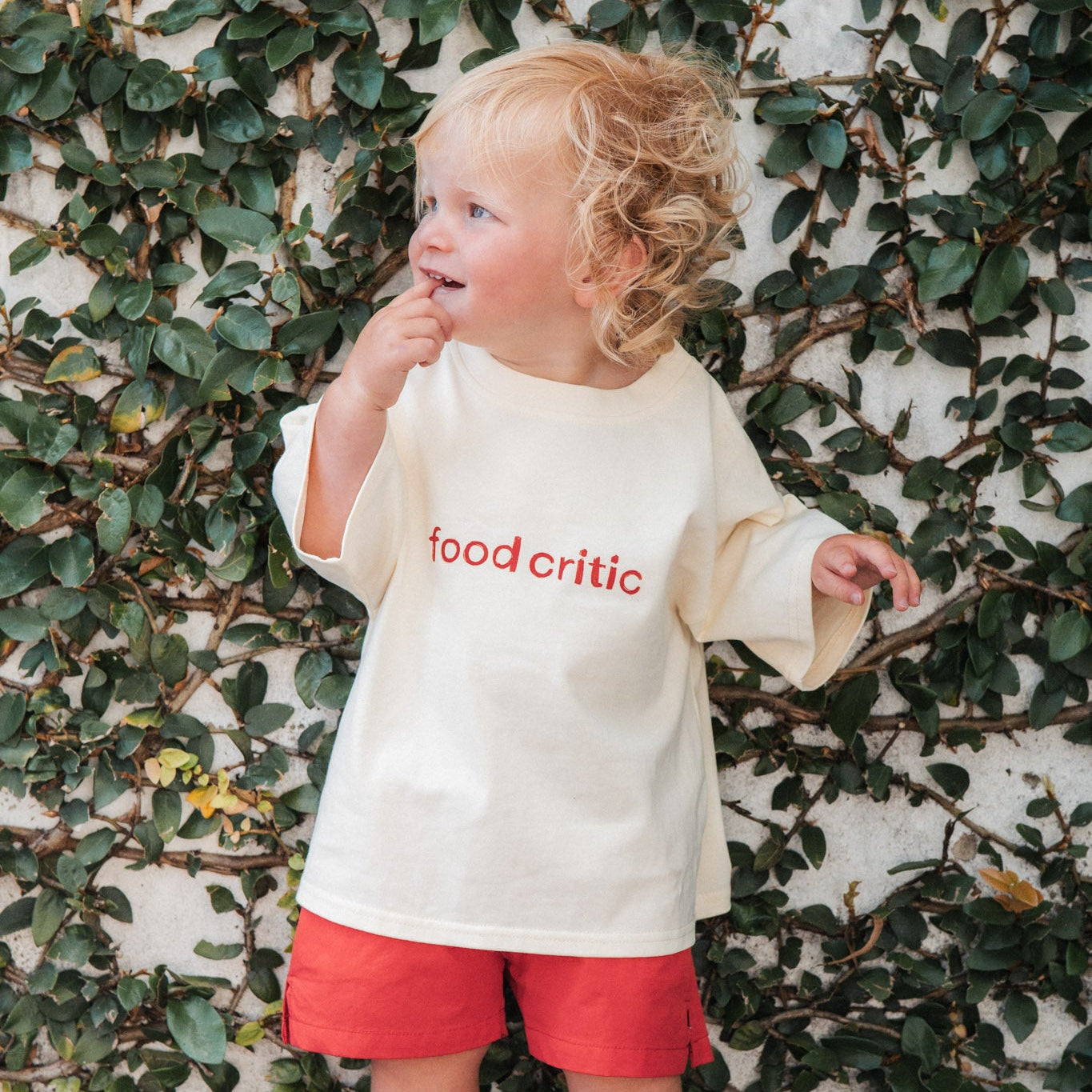Child wearing a 'food critic' shirt in front of a leafy wall