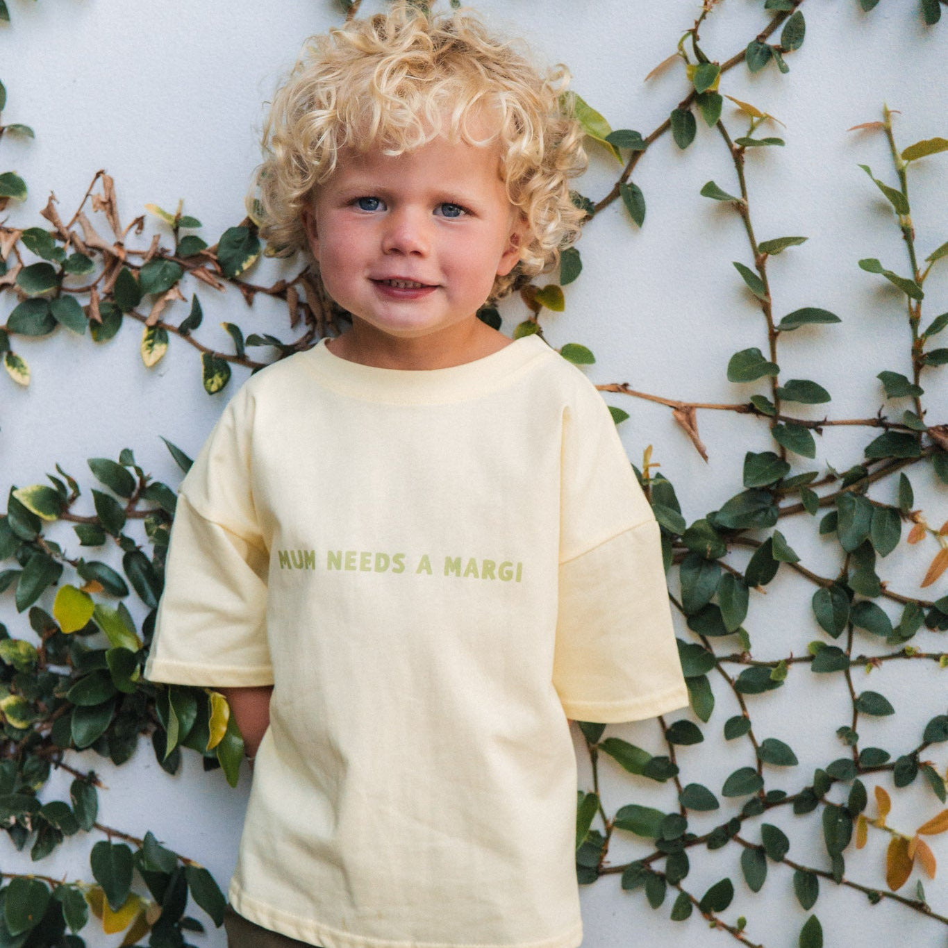 Child wearing a lemon tee with mum needs a margi text, standing against a white wall with green foliage.