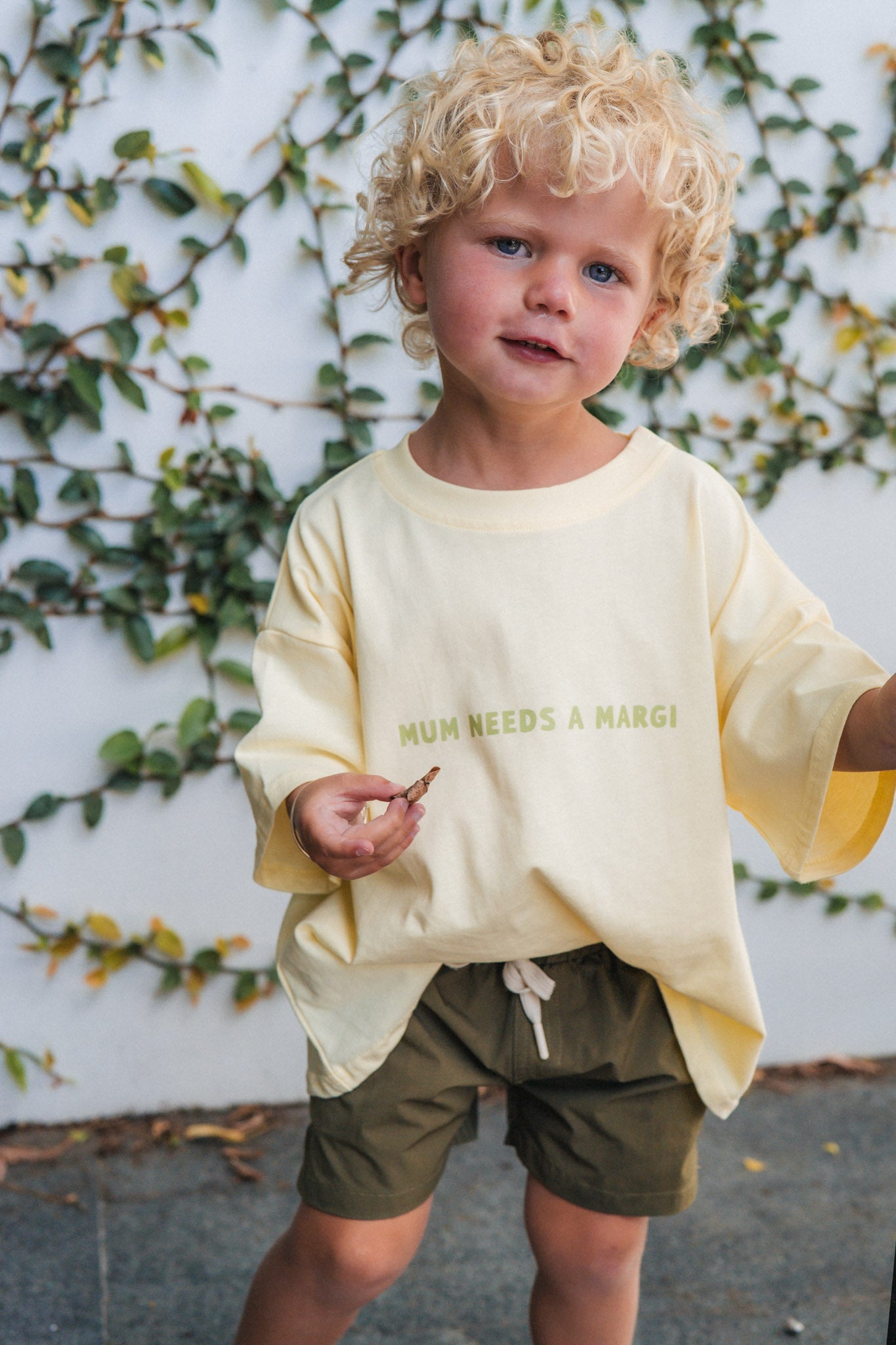 Child wearing a light yellow shirt with text and green shorts standing against a white wall with greenery.