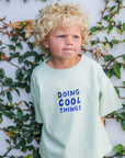 Child wearing a shirt with 'Doing Cool Things' text against a leafy background