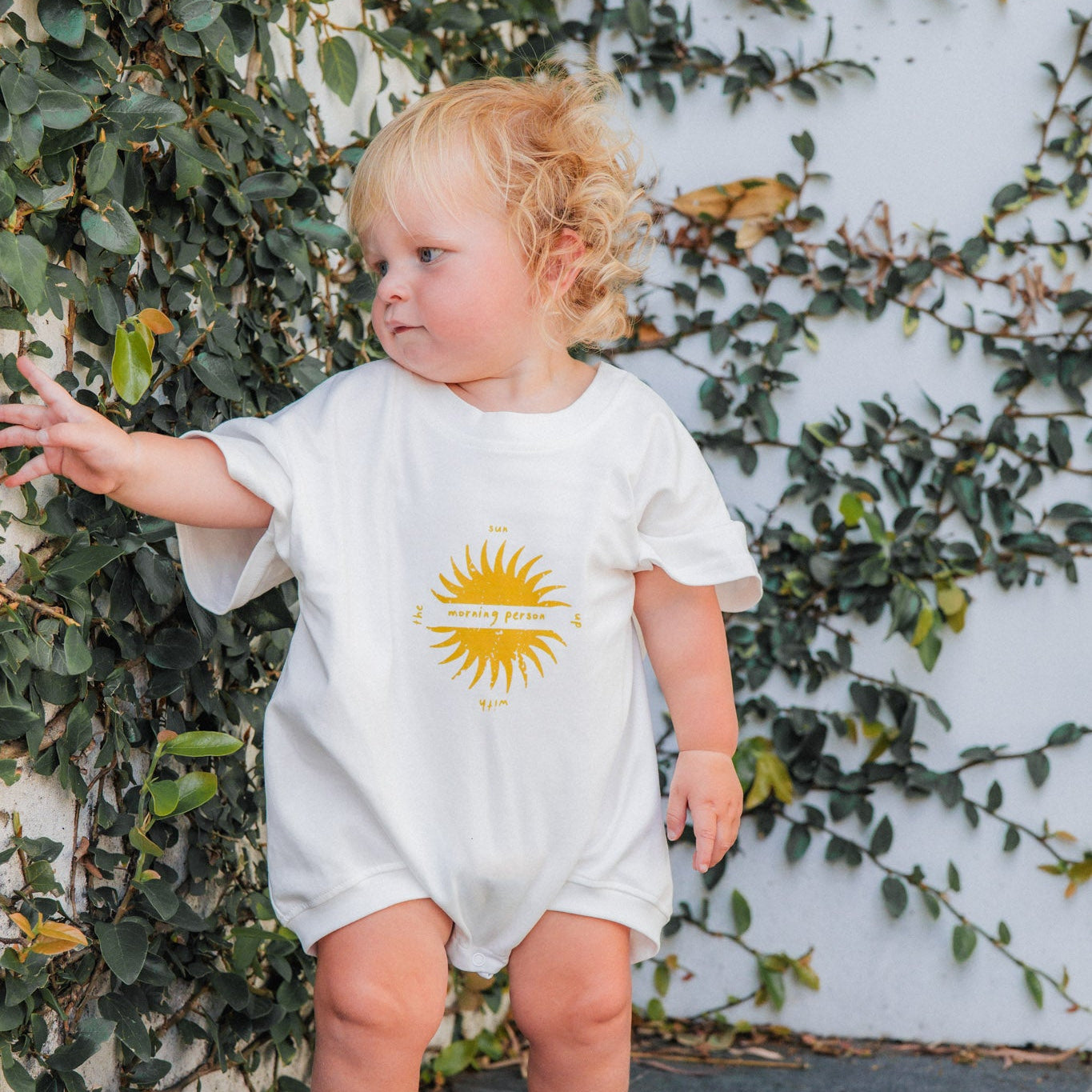 Child wearing a white onesie with a morning person design, standing against a wall with greenery.