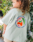 Child wearing family is my world light green shirt with a colorful graphic and text outdoors.