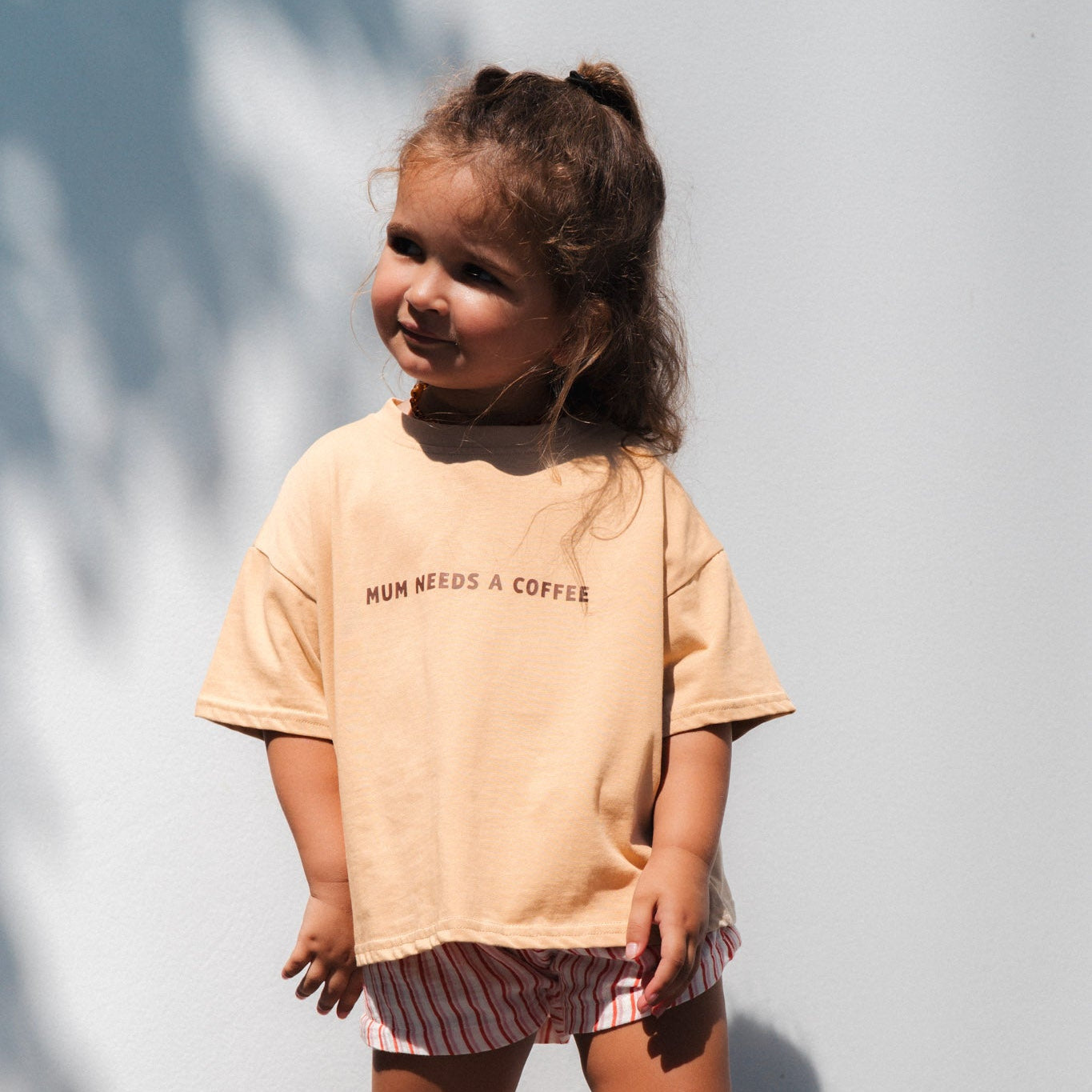 Child wearing a beige t-shirt with mum needs a coffee text and striped shorts standing against a light background