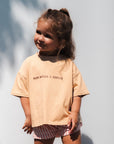 Child wearing a beige t-shirt with mum needs a coffee text and striped shorts standing against a light background