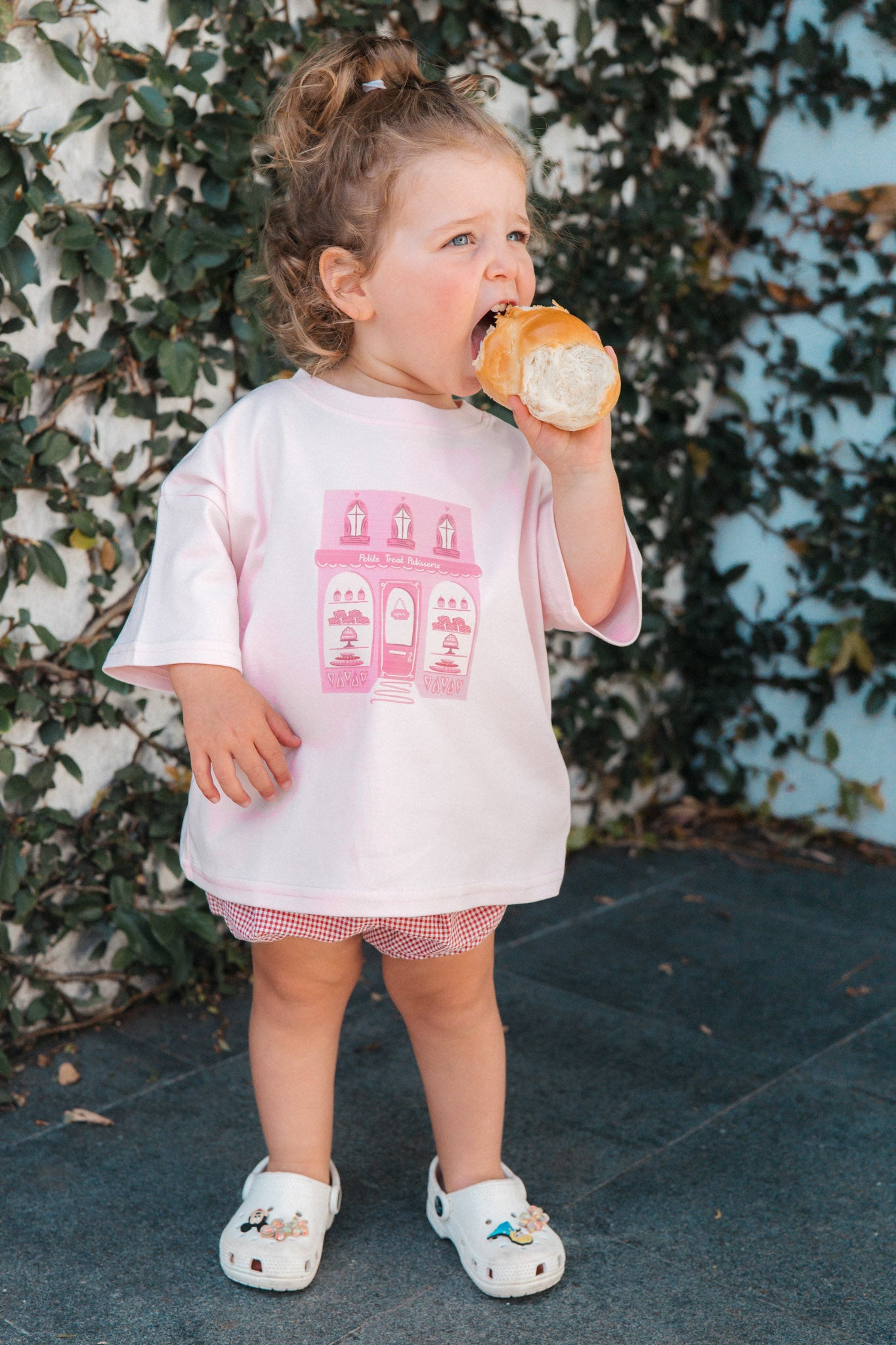 Child eating a donut wearing a pink tee with pink graphics against a leafy background
