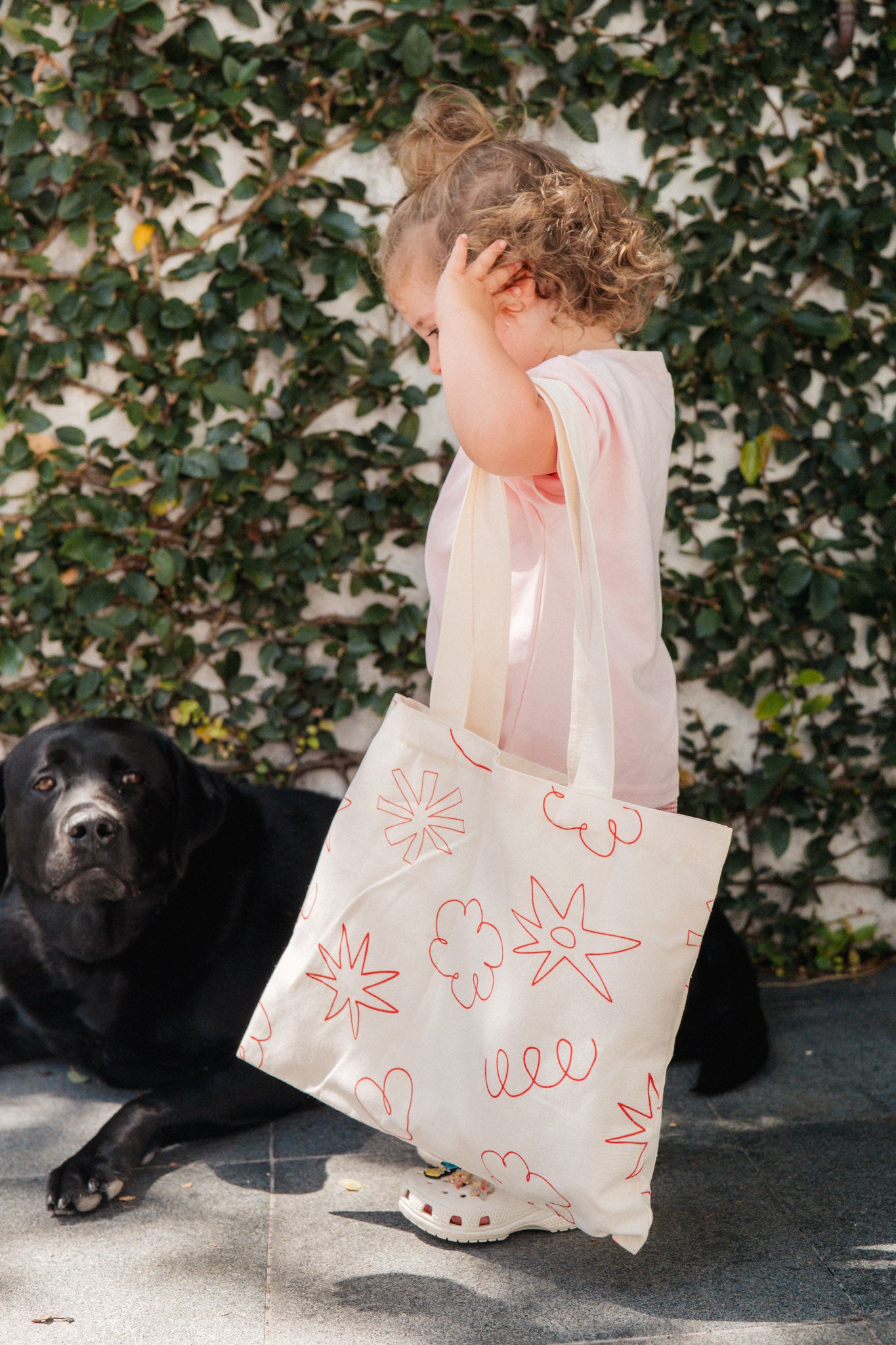 Child holding a tote bag with floral designs next to a black dog against a green leafy background